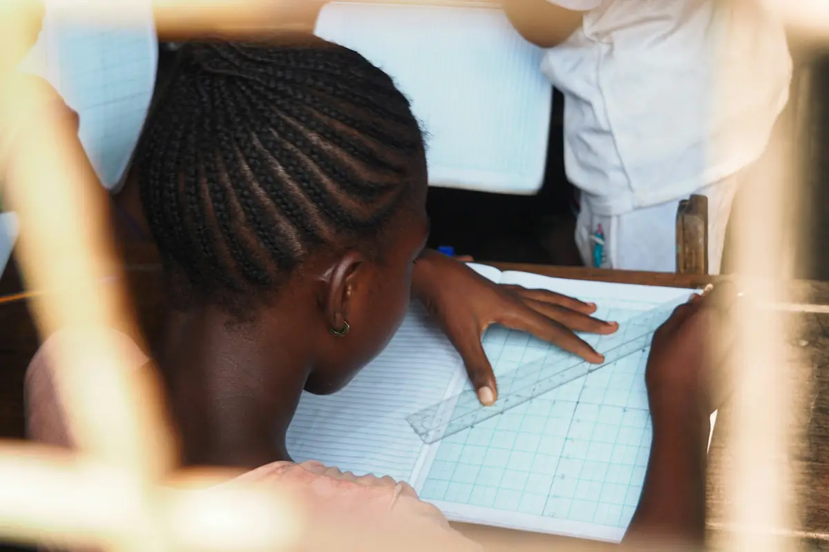 A student, seen from behind, rules lines in a squared exercise book at a wooden school desk